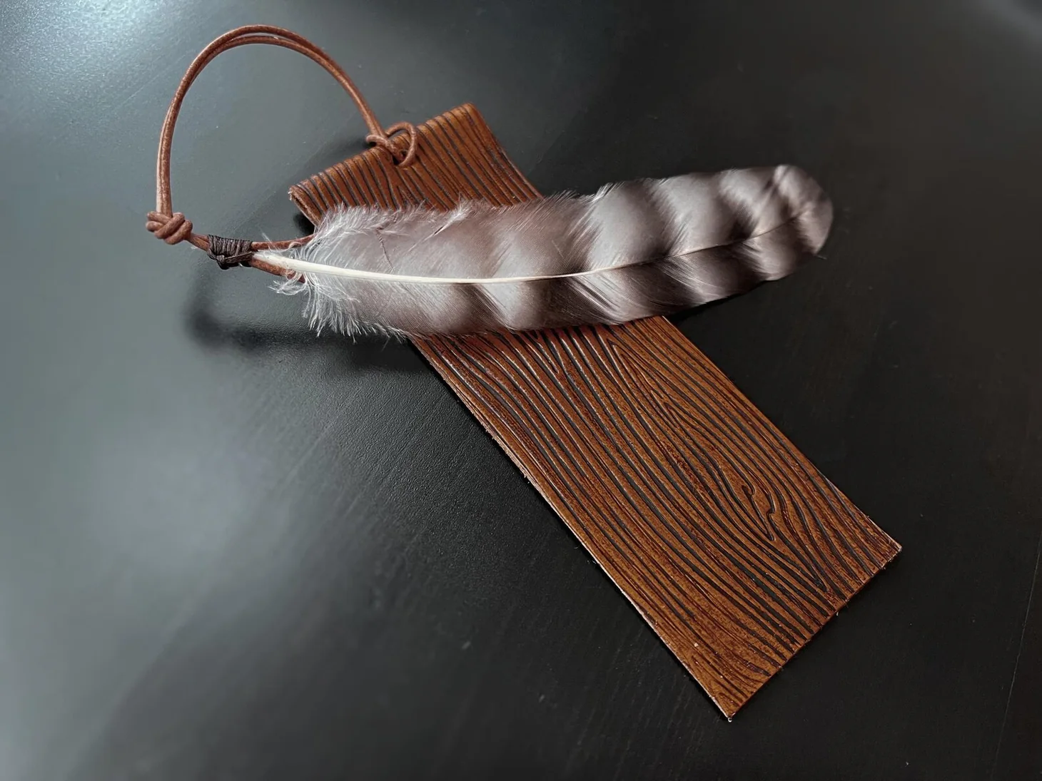 Handmade brown vegetable-tanned leather bookmark with stitched texture and a feather attached by leather cord.